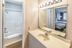 Bathroom with a beige vanity, large mirror, and bright overhead lights, featuring an adjacent room with a bathtub and a view into a bedroom with a neatly made bed.