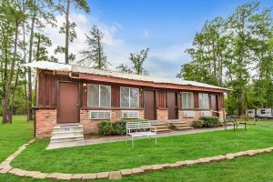 Single-story brick and wood cabin building with multiple units, each having its own door and window air conditioner. The building is surrounded by tall pine trees, with a bench and outdoor seating in front on a small grassy area. An RV is visible in the background.