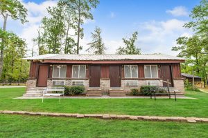 Small rustic motel-style building with four units, brick and wood exterior, white metal roof, front steps, benches, and surrounded by tall trees.