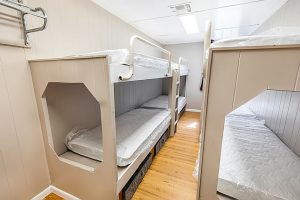 Narrow cabin room with two sets of built-in bunk beds, each with two mattresses wrapped in plastic. The beds have safety rails on the top bunks and wood paneling around the frames. The room has light-colored paneled walls, wood flooring, and a ceiling light.