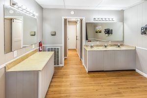 Clean RV park restroom with wood-style flooring, light gray paneled walls, large mirrors above beige countertops, multiple sinks, and bright vanity lighting.