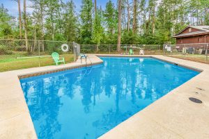 Outdoor swimming pool surrounded by a concrete deck and chain-link fence, with green and white lounge chairs, set against a backdrop of tall pine trees and a rustic building.