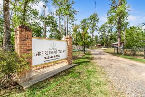 Entrance to Bundick Lake Retreat and RV featuring a large sign with contact information, flanked by brick pillars and surrounded by tall pine trees and a gravel driveway.