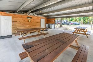 Covered outdoor pavilion with wooden picnic tables on a concrete floor, adjacent to a wood-paneled building with restrooms, overlooking RVs and parked vehicles in a wooded area