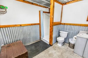 Rustic RV park bathroom with corrugated metal walls, wood framing, a shower with blue curtain, toilet, small sink, and trash can.