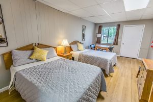 Cozy cabin-style room with two double beds covered in gray quilted bedding and yellow accent pillows. A wooden nightstand with a lamp sits between the beds. The room has wood flooring, a dresser, a small table with chairs near the window, and wall-mounted decor. Natural light comes in through a window with brown curtains.