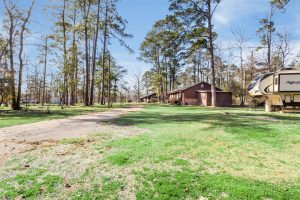 Bundick Lake RV Park Bundick Lake Retreat and RV Park in DeRidder, Louisiana: grassy yard, tall trees, gravel driveway, RV, cabins under a blue sky.